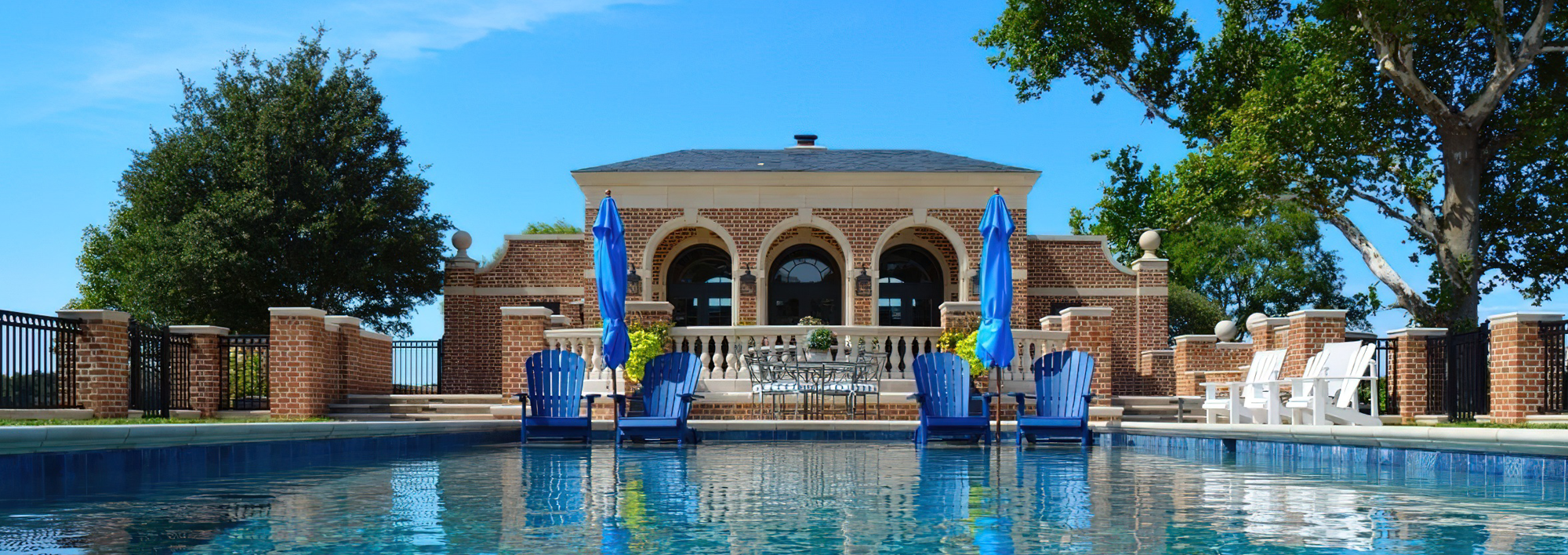 Aqua Pools A view from a pool designed by expert pool sales & design professionals, facing a brick building with arched windows, blue umbrellas, blue chairs, white chairs, and green trees under a clear blue sky.