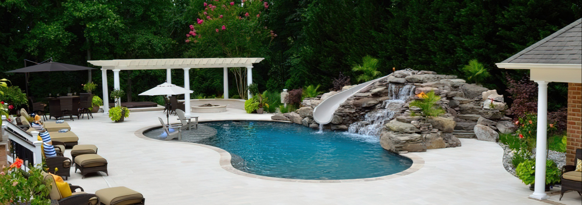 Aqua Pools A backyard pool with a rock waterfall, water slide, lounge chairs, umbrella, pergola, and lush greenery surrounding the area—an inviting oasis that would make a stunning addition to any portfolio.