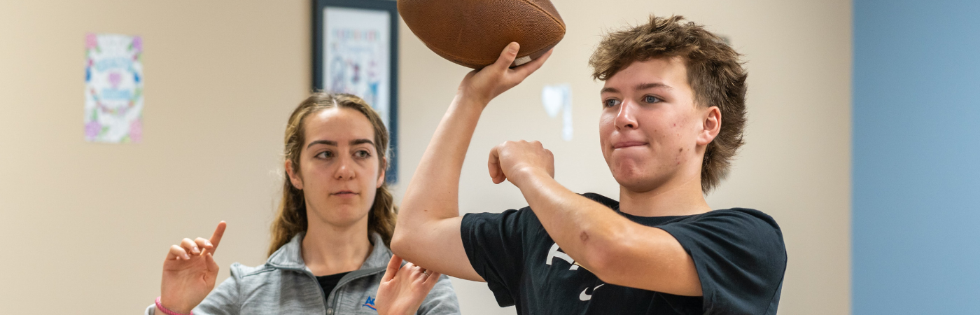 a young man holding a football while being instructed by a female physical therapist.