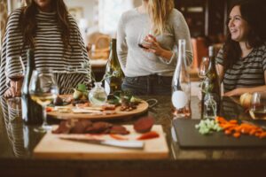 three women standing around a table with food and wine