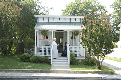 The Atlantic Hotel Victorian Gardner’s Cottage two women dressed up in victorian dresses on front porch of small white cottage surrounded by trees on a sunny day