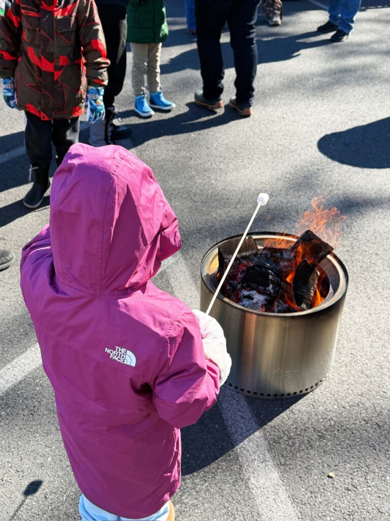 a little girl in a purple jacket standing next to a pot of food