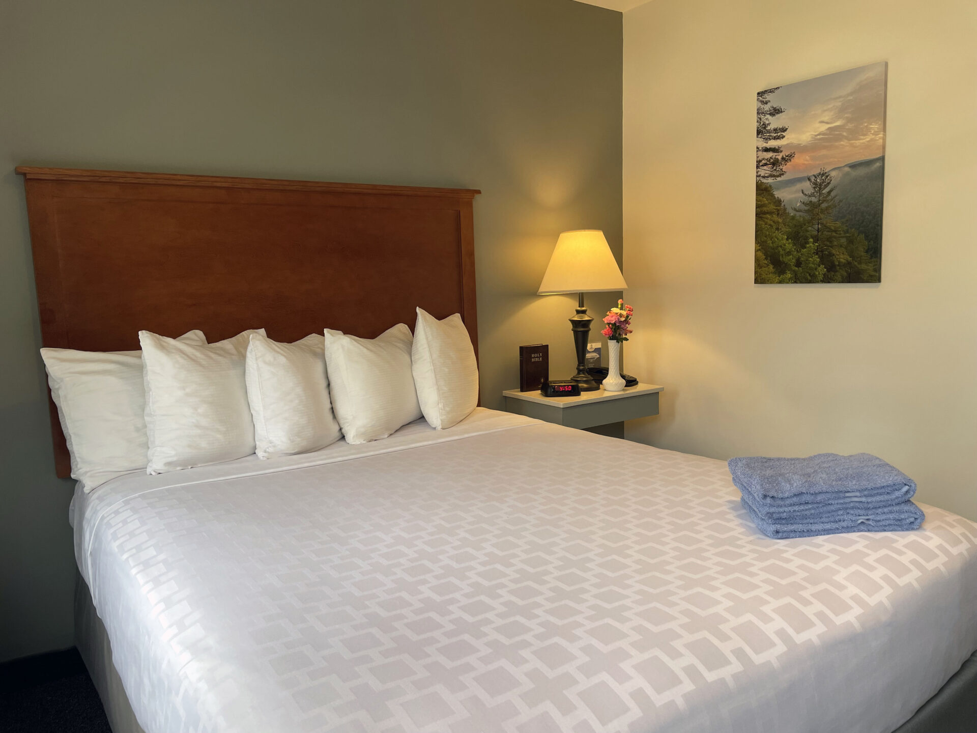 Bed made with white bed sheets and a stack of blue towels with a painting hanging on the wall