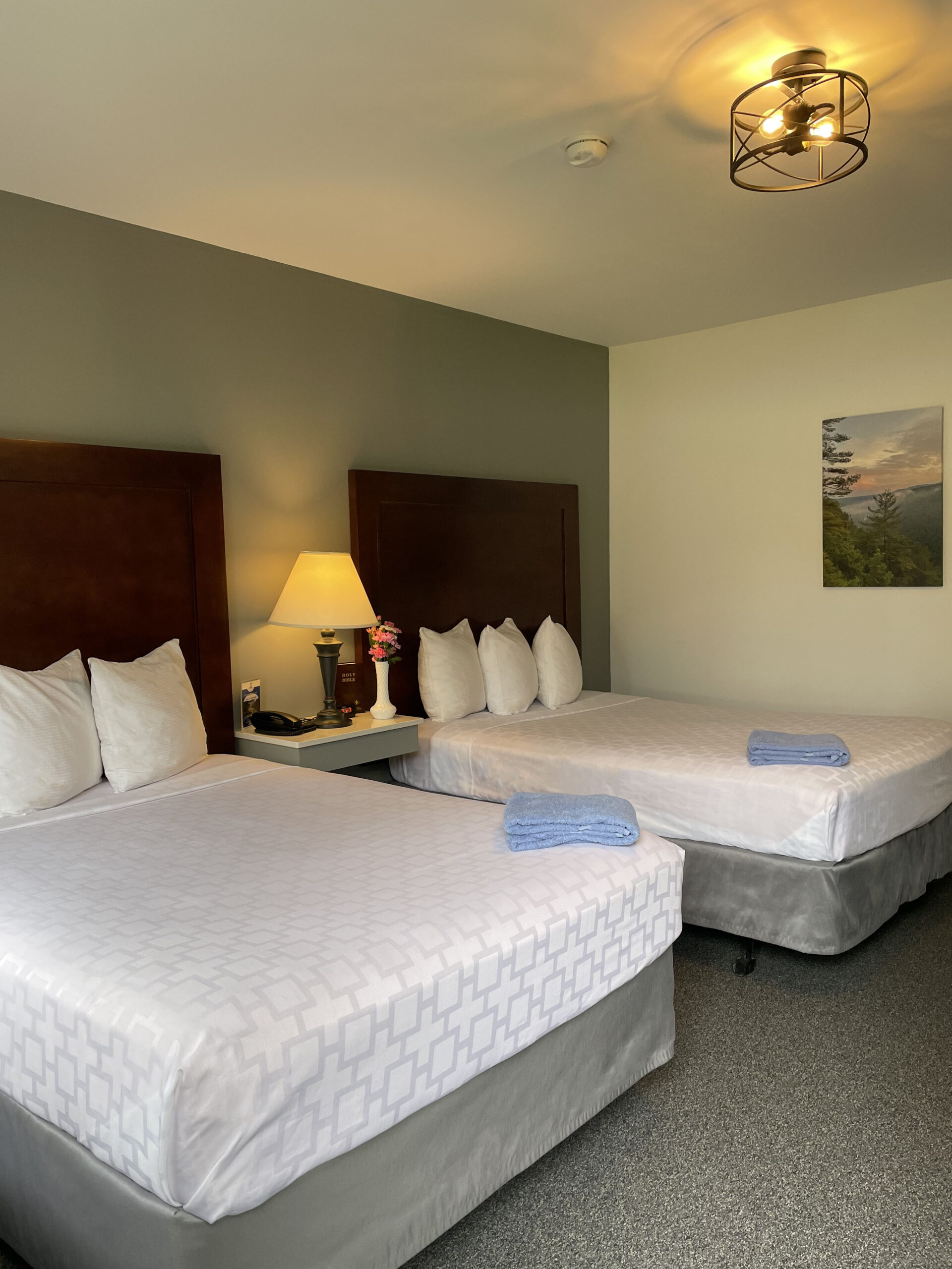 Two beds made, with blue towels at the foot and a nightstand with a lamp between them