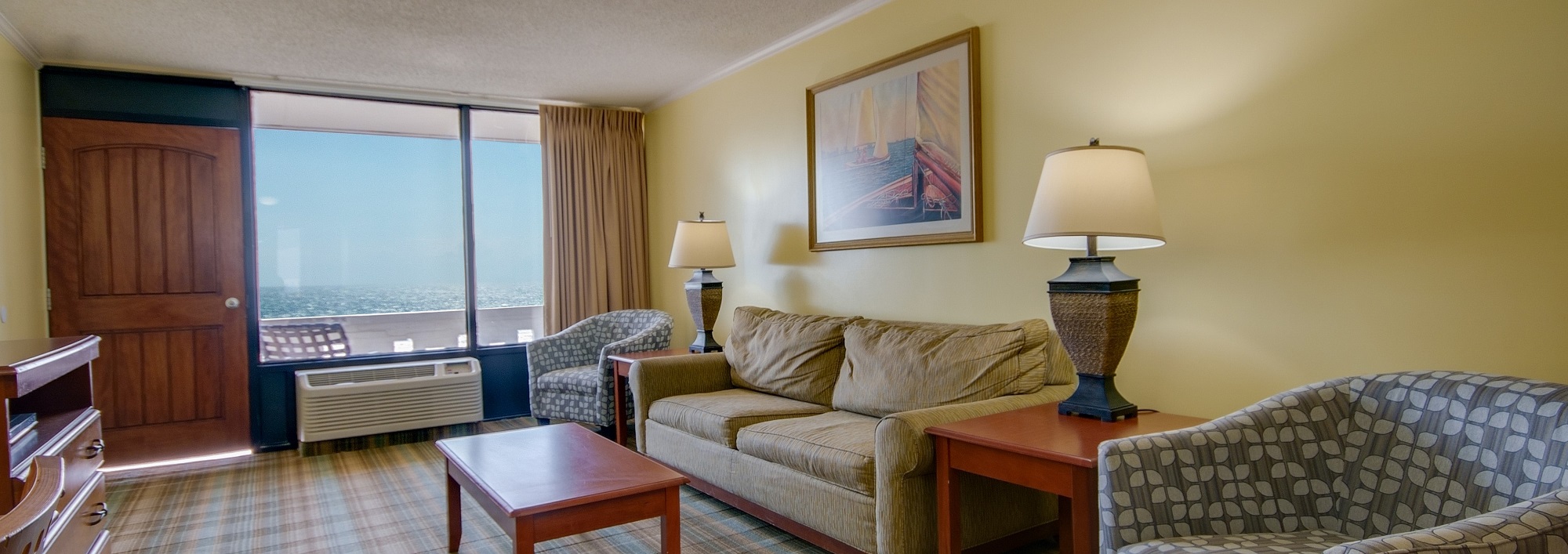 A living room with a sofa, two armchairs, a coffee table, and lamps, with a large window showing an ocean view.
