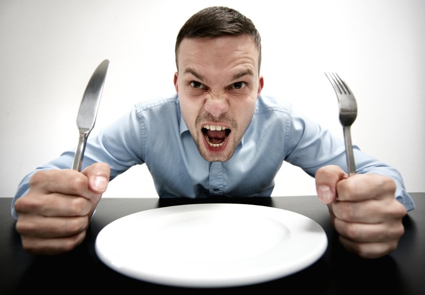 Man holds fork and knife with empty plate and angry look on face