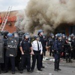 Police personnel line up protected with shields in front of CVS building with smoke in background