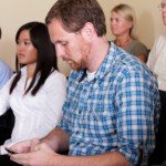 Man in blue plaid shirt sits among others while texting on cell phone