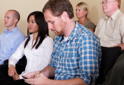 Man in blue plaid shirt sits among others while texting on cell phone