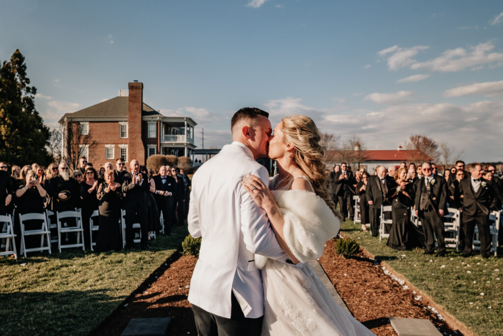 A bride and groom kissing with guests in the background at the outdoor ceremony space at King Cole Farm.