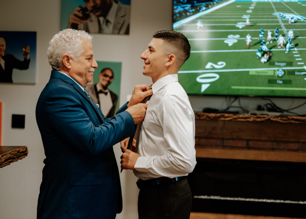 A groom and his father putting on a tie in the groomsmen's lodge in the pole barn at king cole farm.