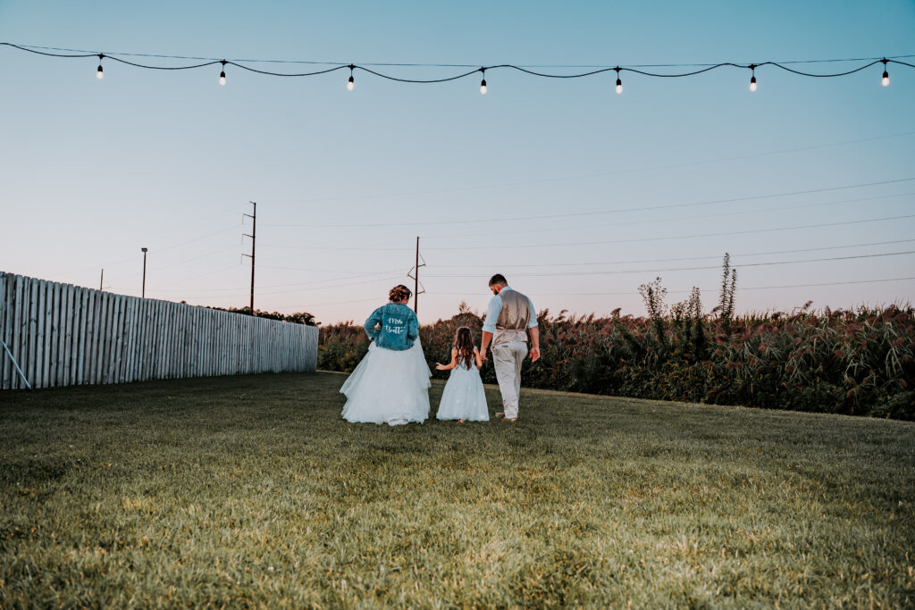 A bride and groom and flower girl against the corn field in the back patio of the Pole barn at King Cole Farm.