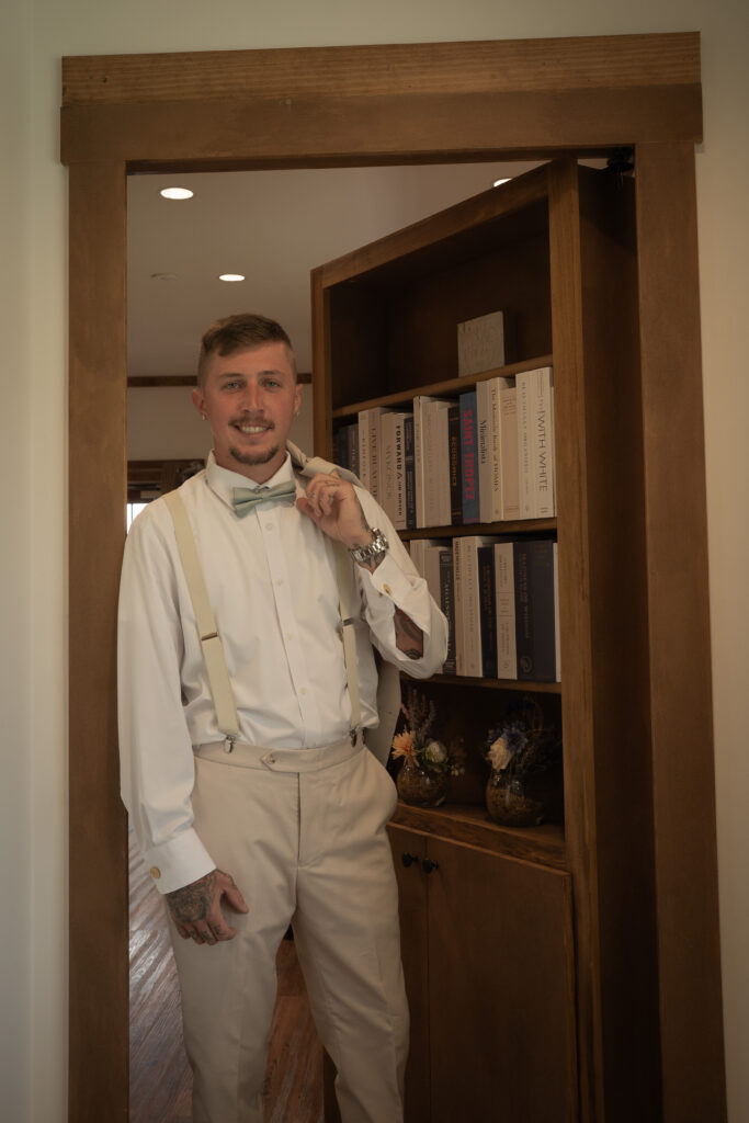 A groom leaning against a doorframe wiht a bookcase door behind him outside of the groomsmen's lodge in the Pole Barn at King Cole Farm.