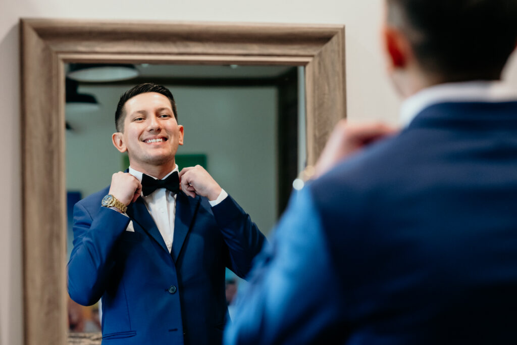 A groom adjusting his bow tie in the mirror in the Groomsmen's Lodge at King Cole Farm.