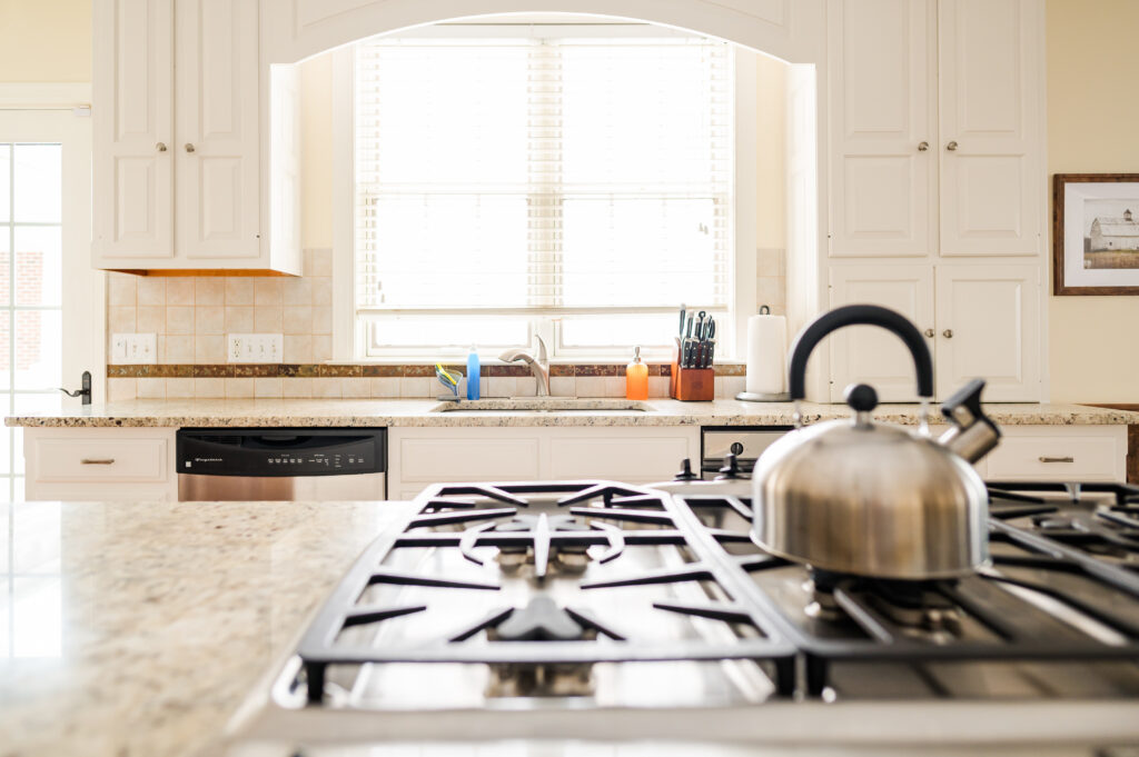 Image including the kitchen countertops, kitchen island with range, tea kettle, sink, dishwasher, and windows in the Residence at King Cole Farm.