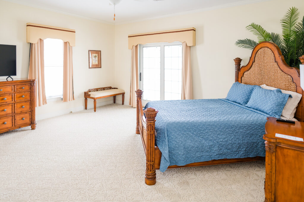 Image featuring a bed, nightstand with a lamp, dresser with tv, window and sliding glass door, bench and decorative palm tree in the Island Sun Bedroom at King Cole Farm