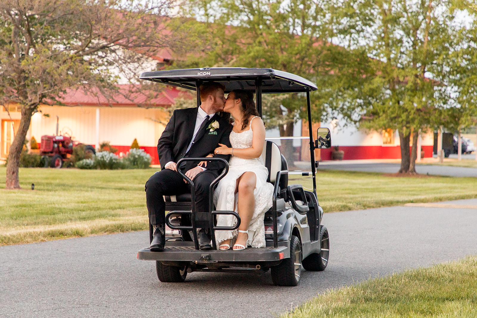 A Bride and Groom driving away in the getaway golf cart at King Cole Farm with the Pole Barn reception space in the background.