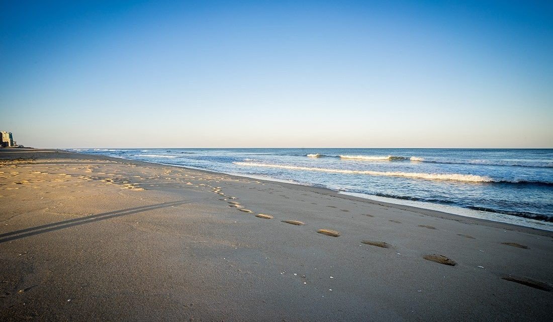 A serene beach scene with footprints in the sand, waves, and a clear blue sky.