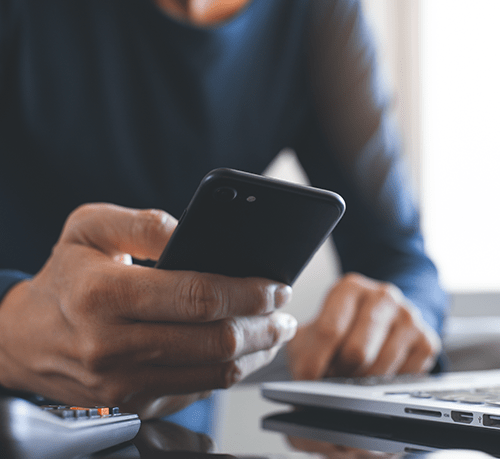 close of man's hand holding cell phone with laptop