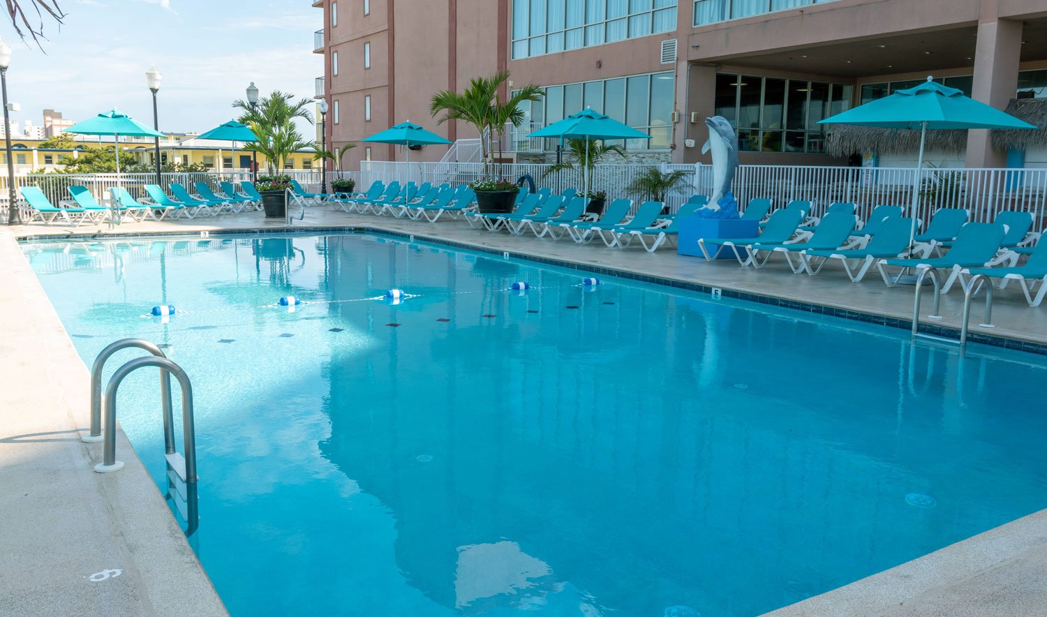 A large outdoor pool with blue lounge chairs and teal umbrellas, surrounded by palm trees and a dolphin statue, in front of a multi-story building.