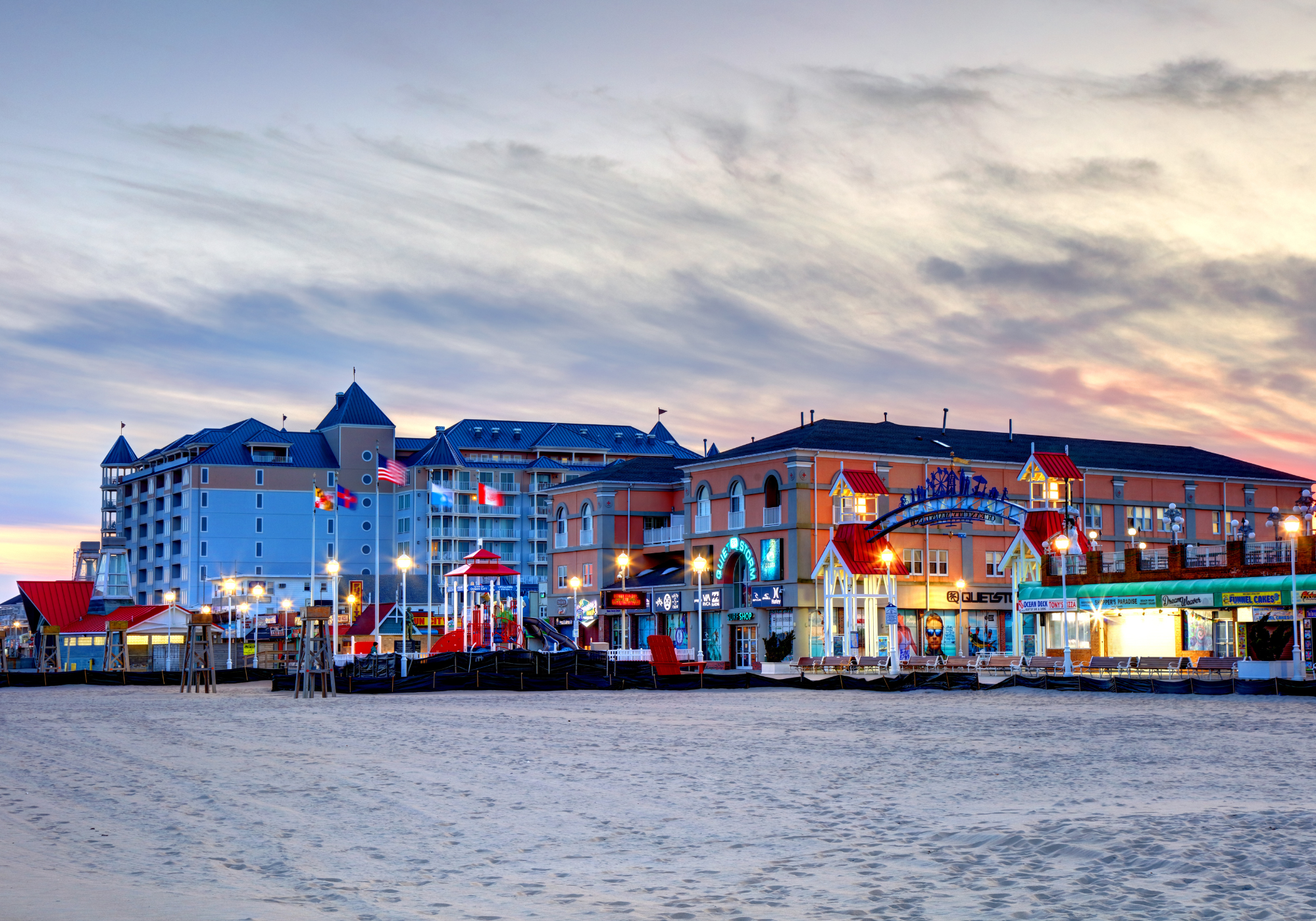 A wide view of a beachfront hotel at sunset, with a sandy beach in the foreground and a multi-story building in the background.