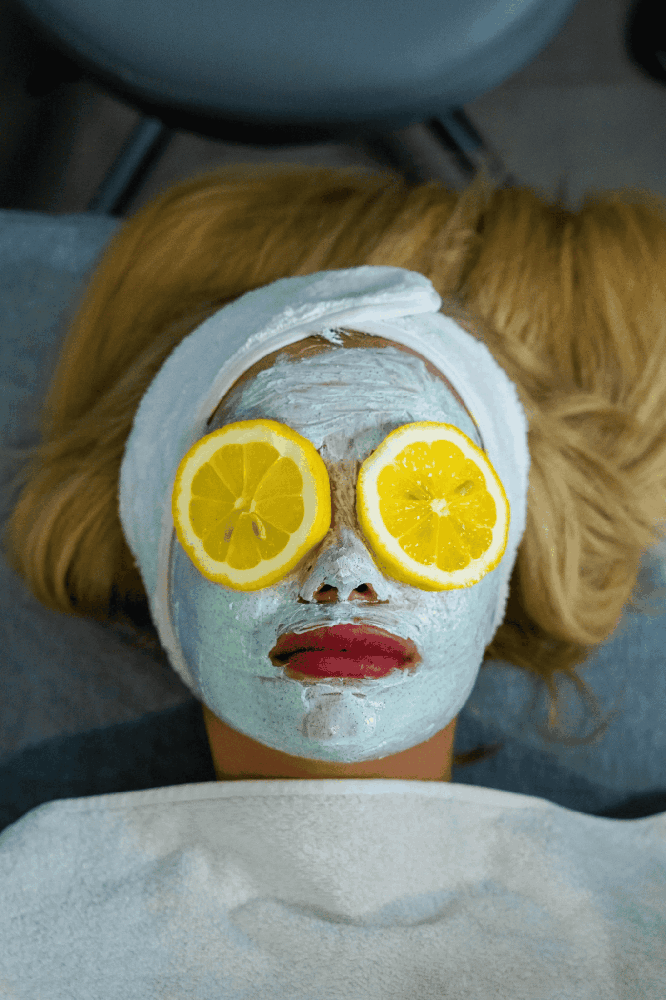 A woman receiving a facial treatment with a blue mask and lemon slices on her eyes.