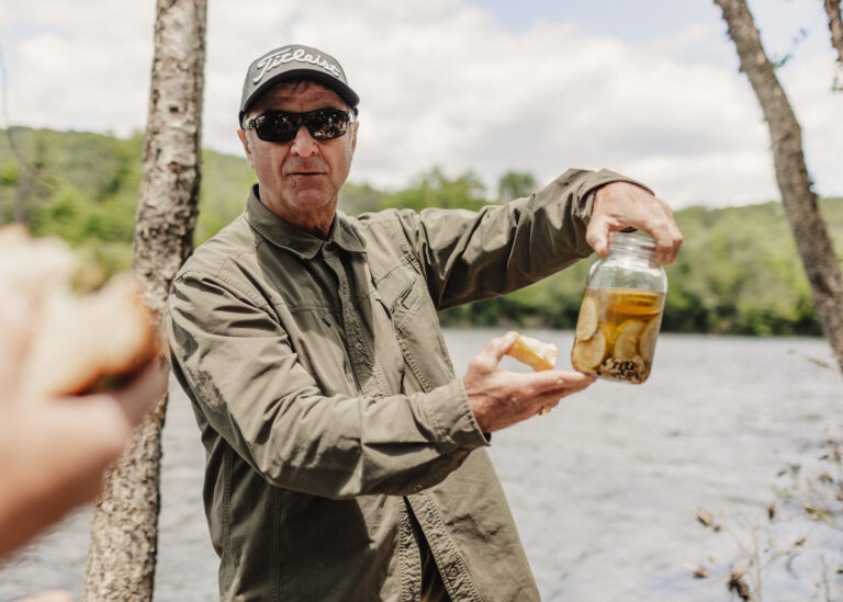 a man holding a jar of beer next to a river